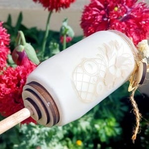 Op de afbeelding: Een witte flesstopper met een ananasontwerp, met een houten basis en een houten stok. De stopper is versierd met een jute touw en kleine witte bloemen. De achtergrond bevat rode bloemen en groen gebladerte.