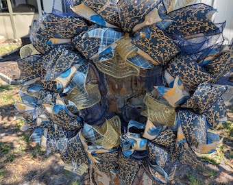 Könnte beinhalten: Ein dekorativer Kranz mit einem Netzsockel und geschichteten Bändern. Die Bänder weisen Muster in Marineblau, Gold und Hellblau auf. Der Kranz wird im Freien an einem Baumstamm befestigt.