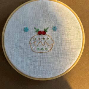 Puede incluir: Un aro de bordado blanco con un diseño de punto de cruz de un pudín navideño con bayas rojas y hojas de acebo verdes. Hay dos copos de nieve azules sobre el pudín.