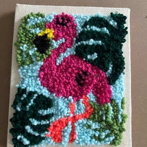 May include: A pink flamingo with a yellow beak and black eyes is surrounded by green and blue leaves. The flamingo is standing on a red flower. The artwork is made with yarn and is displayed on a canvas.