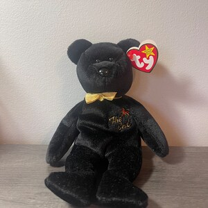 Può includere: Orso di peluche nero Beanie Baby con un fiocco di raso dorato e un'etichetta rossa e dorata "The End". L'orso ha un'etichetta a forma di cuore rossa Ty con una stella bianca e le parole "Beanie Baby" in bianco.