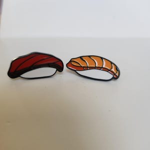May include: Two sushi-shaped pins. One pin features a red topping, the other has an orange topping. Both have a white base and black outline. The pins are on a white surface.