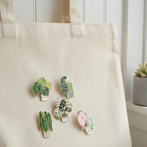May include: A collection of enamel pins featuring various potted plants, including Monstera and other leafy varieties. The pins are in shades of green, white, and pink, and are attached to a beige tote bag. The background includes a white wall and a glimpse of a succulent.