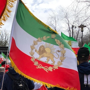 Puede incluir: Bandera iraní con franjas horizontales verdes, blancas y rojas. En el centro, un emblema dorado que representa un león y un sol. Flecos dorados en el borde superior. Otras banderas son visibles en el fondo.