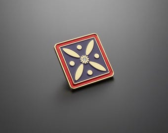 May include: Square-shaped pin with a gold border and a red and blue design. The pin features a gold floral pattern with a small, clear gem in the center. The pin is likely made of metal and has a shiny finish.