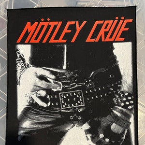 May include: Black and white image of a person wearing black leather pants and a studded belt. The text "MÖTLEY CRÜE" is in red at the top of the image. The text "© 2004 Mötley Crüe Touring, Inc." is at the bottom of the image.