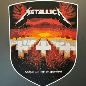 Peut inclure: Un autocollant noir et blanc en forme de bouclier. L'autocollant présente le logo du groupe Metallica et le titre de l'album "Master of Puppets". L'image montre un coucher de soleil avec un champ de croix et des mains tenant des cordes.