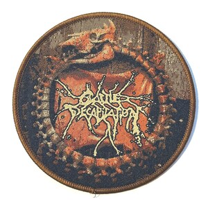 Op de afbeelding: Ronde patch met een bruine rand en een gedetailleerd ontwerp. Het midden toont de tekst "DECAPITATION" in een gestileerd lettertype, met een schedel en andere grafische elementen in bruin- en oranje tinten.