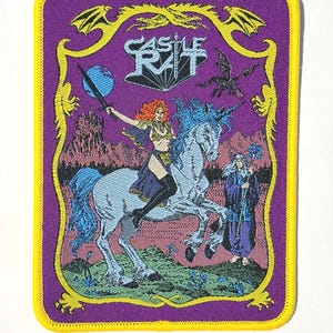 Op de afbeelding: Een rechthoekige geborduurde patch met een gele rand op een paarse achtergrond. Het toont een figuur met rood haar op een lichtblauwe eenhoorn, die een zwaard naar een blauwe bol houdt. Een figuur in een gewaad staat ernaast, met de tekst CASTLE RAT.