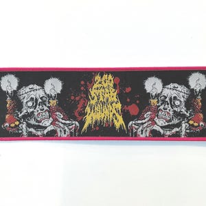 May include: A rectangular patch with a black background and a pink border. The patch features two zombie-like figures with candles, and a central yellow and red graphic. The text on the patch is illegible.