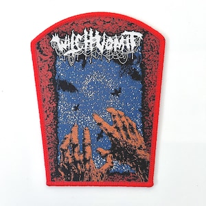 May include: Embroidered patch with a red border. The design features the text "Witch Vomit" in white, above a blue background with orange hands reaching upwards. The patch has a black and red floral design.