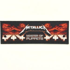 Puede incluir: Parche tejido negro y rojo con el texto "Metallica Master of Puppets" y un gráfico rojo y naranja de una mano que se extiende desde un fondo oscuro.