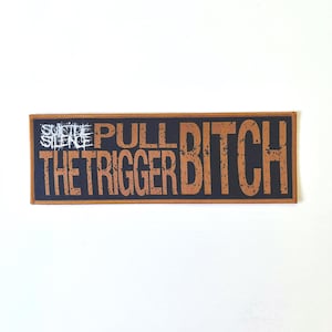 May include: A rectangular patch with the words "SUICIDE SILENCE" in white and "PULL THE TRIGGER BITCH" in brown. The patch has a black background and a brown border.