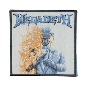 Puede incluir: Parche bordado con el nombre de la banda "MEGADETH" en azul, sobre una ilustración de una figura esquelética con traje, con llamas. El parche tiene un borde negro y un fondo claro.