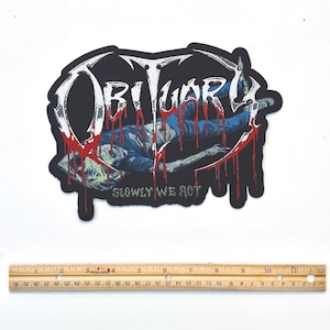 Può includere: Toppa nera con la scritta "OBITUARY" in bianco, con sangue rosso che gocciola. Sotto è raffigurata una figura in blu, con la scritta "SLOWLY WE ROT". La toppa è larga circa 13 cm.