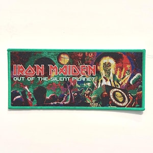 以下が含まれることがあります： 赤い文字と白い文字で「Iron Maiden」と「Out of the Silent Planet」と書かれた緑色の刺繍パッチ。パッチには、赤と黒の衣装を着た人物が写った、カラフルなコンサートシーンの画像が描かれている。