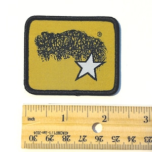 May include: A rectangular embroidered patch with a mustard yellow background and a black border. It features a black graphic of a shaggy creature and a white five-pointed star. A wooden ruler shows the patch is approximately 6.35 cm wide.