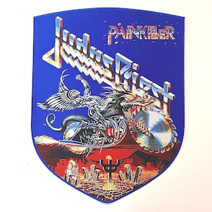 Puede incluir: Parche azul en forma de escudo con el nombre de la banda "Judas Priest" en una fuente multicolor y dentada, con la palabra "Painkiller" encima. La imagen representa un jinete alado en una motocicleta con una rueda de hoja de sierra, sobre un paisaje urbano.