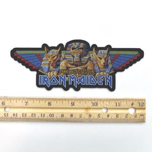 May include: An embroidered Iron Maiden Powerslave patch featuring an Egyptian-themed graphic with a central pharaoh figure flanked by two jackal heads and wings. A wooden ruler placed below shows the patch is approximately 6 inches wide.