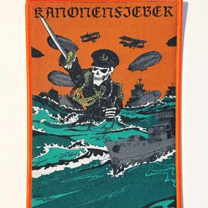 Puede incluir: Un parche bordado rectangular con borde naranja que muestra un esqueleto con uniforme militar sosteniendo una espada. El fondo muestra un mar con buques de guerra, aviones y dirigibles. El texto KANONENFIEBER aparece en la parte superior.