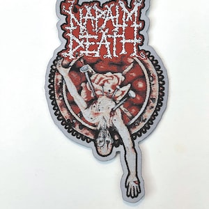 Puede incluir: Un parche bordado blanco y rojo con el nombre de la banda "NAPALM DEATH" en letras blancas. El parche presenta un gráfico de una persona clavada a una rueda.