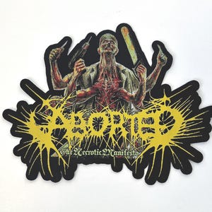 Pode incluir: Um remendo bordado preto e amarelo com a palavra "ABORTED" em um design de respingo. O texto "The Neurotic Manifesto" está abaixo da palavra "ABORTED". O remendo apresenta uma imagem gráfica de uma pessoa com vários braços segurando várias ferramentas.