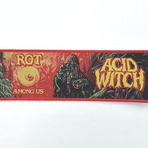 May include: A rectangular patch with a red border and a dark, detailed graphic. The patch features the words "ROT AMONG US" and "ACID WITCH" in yellow lettering. The artwork depicts a hooded figure and a large eye.