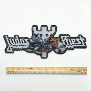 Puede incluir: Parche bordado con el nombre de la banda "Judas Priest". El parche presenta un gráfico detallado de una criatura alada montando una motocicleta con una rueda de hoja de sierra. El parche mide aproximadamente 30 cm de largo.