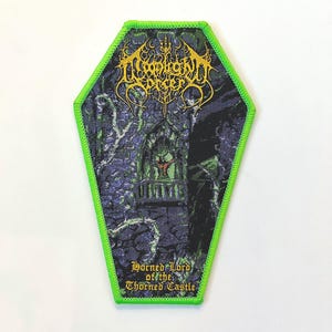 May include: A green and black embroidered patch with a coffin shape. The patch features a band logo with the text "Horrendous" in gold lettering. The text "Horned Lord of the Thorned Castle" is also in gold lettering.