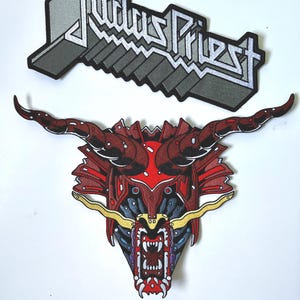May include: Two embroidered patches. The top patch features the band name "Judas Priest" in a gray, 3D-style font. Below is a red and black dragon head patch with horns and sharp teeth. The dragon's mouth is open, revealing white teeth.