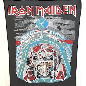 Puede incluir: Un parche de banda Iron Maiden negro con un logotipo rojo y blanco. El parche presenta un gráfico de un piloto con casco y gafas, con una mira de objetivo delante de la cara del piloto. El piloto está en una cabina con un avión azul y verde en el fondo.