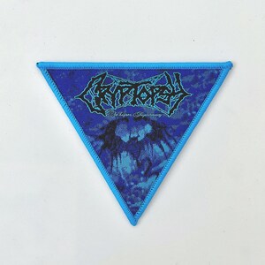 May include: A blue triangular patch with a black and white band logo that says "Cryptopsy" and the text "Whisper Supremacy" in white. The patch has a blue border.