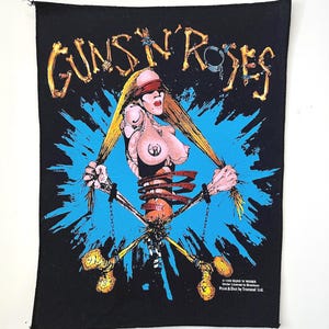May include: A black fabric patch featuring a Guns N' Roses graphic. The design shows a figure with long blonde hair, a red blindfold, and chains attached to wrists. The background has a blue splash effect, and the text reads Guns N' Roses.