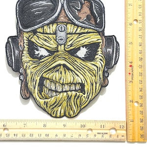 Puede incluir: Parche bordado con un rostro esquelético amarillo con casco y gafas de piloto. El parche tiene detalles en negro, marrón y amarillo. El parche mide aproximadamente 10 cm de ancho y 10 cm de alto.