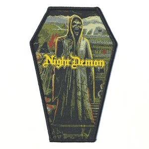 May include: A coffin-shaped patch featuring a hooded figure with a skeletal face, set against a backdrop of a building and landscape. The patch has the words "Night Demon" in yellow, gothic-style lettering.