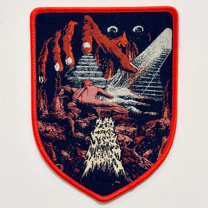 May include: A red, white, and black embroidered patch with a detailed image of a hand reaching out from a dark background. The hand is holding a white object with a red outline. The patch has a red border and a pointed shape.
