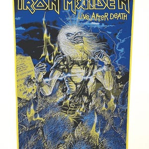 Könnte beinhalten: Ein blauer und gelber Iron Maiden Patch mit dem Bandnamen und den Worten "Live After Death" in Gelb. Der Patch zeigt ein Schwarzweißbild eines Mannes mit langen Haaren und einer Kette um das Handgelenk. Der Mann steht in einem Grasfeld mit Flammen hinter ihm.