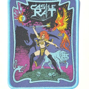 Op de afbeelding: Een rechthoekige geborduurde patch met een lichtblauwe rand en de tekst CASTLE RAT bovenaan. Het ontwerp toont een roodharig figuur met een zwaard, een feniks, een wolf, een zon- en maansymbool en schedels op een paarse achtergrond.