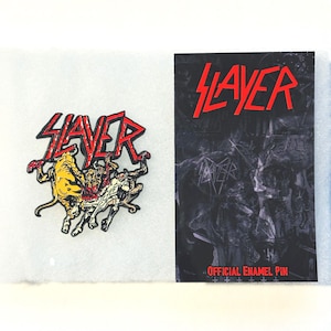 May include: An enamel pin and a card featuring the band Slayer. The pin has a red Slayer logo above a graphic of figures. The card has the Slayer logo in red and the words "OFFICIAL ENAMEL PIN".