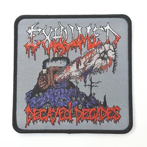 Può includere: Una toppa ricamata grigia con il nome della band "Exhumed" in lettere rosse e bianche e il titolo dell'album "Decayed Decades" in lettere rosse. La toppa presenta una motosega insanguinata e un braccio mozzato.