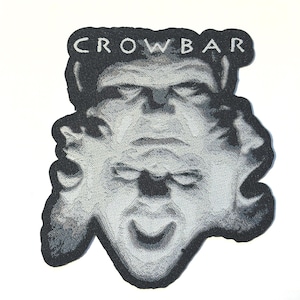 May include: A black and white patch featuring a stylized face with the word "CROWBAR" arched above. The design has a textured appearance, with shading creating depth and dimension. The patch is likely for clothing or accessories.