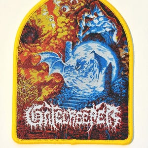 May include: A shield-shaped embroidered patch with a bright yellow border. The design features a blue winged creature perched on a sphere against a fiery orange and red background. The text Gatecreeper is printed in white stylized lettering.