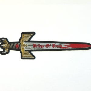 Puede incluir: Parche bordado con forma de espada, con una hoja roja y una empuñadura dorada. El texto "Bridge Of Death" está bordado en rojo sobre la hoja. El parche tiene un borde negro.