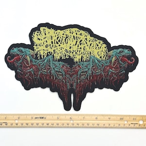 May include: A black fabric patch featuring a symmetrical illustration of two teal and red monstrous creatures with exposed teeth and tongues, topped with yellow illegible text. A wooden ruler shows the patch is approximately 11 inches wide.