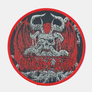 Puede incluir: Parche redondo con borde rojo que presenta una ilustración detallada de una figura demoníaca con cuernos y alas. El diseño es principalmente negro, rojo y blanco, con el texto "HARDCORE DEATH METAL" y "CORPSE GRINDER" en fuente gótica.