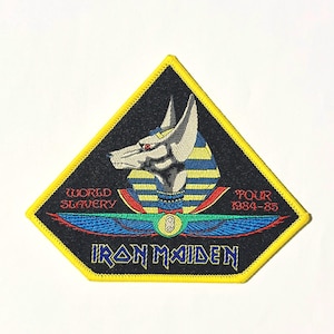 Puede incluir: Un parche triangular negro con borde amarillo. El parche presenta una imagen estilizada de un dios egipcio con alas y el texto "Iron Maiden" en una fuente azul y roja. El texto "World Slavery Tour 1984-85" también está incluido.