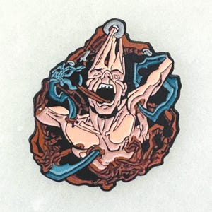 May include: A pin featuring a screaming figure with a wide open mouth, rendered in shades of tan, brown, and blue. The figure appears to be emerging from a textured, brown background, with blue accents and a silver ring at the top.