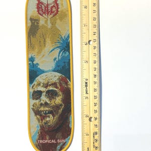 Könnte beinhalten: Ein gestickter Aufnäher mit gelbem Rand. Der Aufnäher zeigt unten ein Zombiegesicht, in der Mitte eine tropische Szene und oben ein rotes Logo. Der Text "TROPICAL SUN" steht unten. Der Aufnäher ist etwa 15 cm lang.