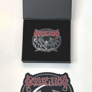 May include: A black and white patch with a red logo for the band Dissection. The logo features a skull with wings and a scythe. The text "Anti-Cosmic Metal of Death" is written on a banner below the skull.