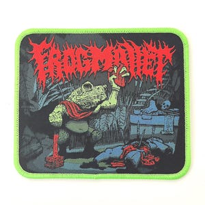 Puede incluir: Un parche rectangular bordado con un borde verde neón brillante. El diseño muestra una criatura parecida a un sapo verde sosteniendo un corazón rojo, con el texto FROG MALLET en una fuente roja dentada arriba. Una figura caída yace abajo.